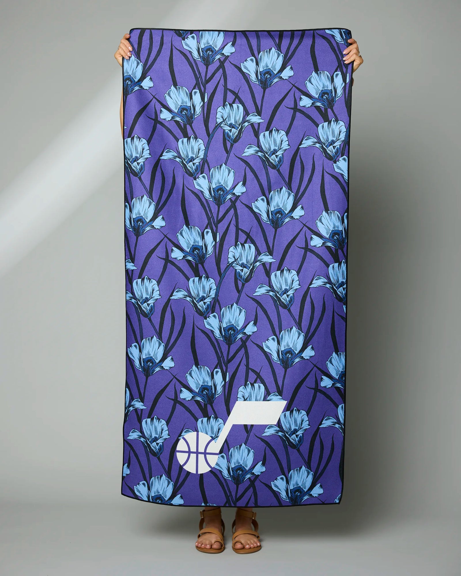 A person holds up a large towel with blue floral patterns on a dark purple background and a white geometric logo in one corner. Only the persons hands and sandals are visible.