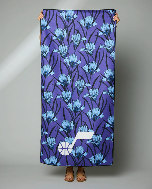 A person holds up a large towel with blue floral patterns on a dark purple background and a white geometric logo in one corner. Only the persons hands and sandals are visible.