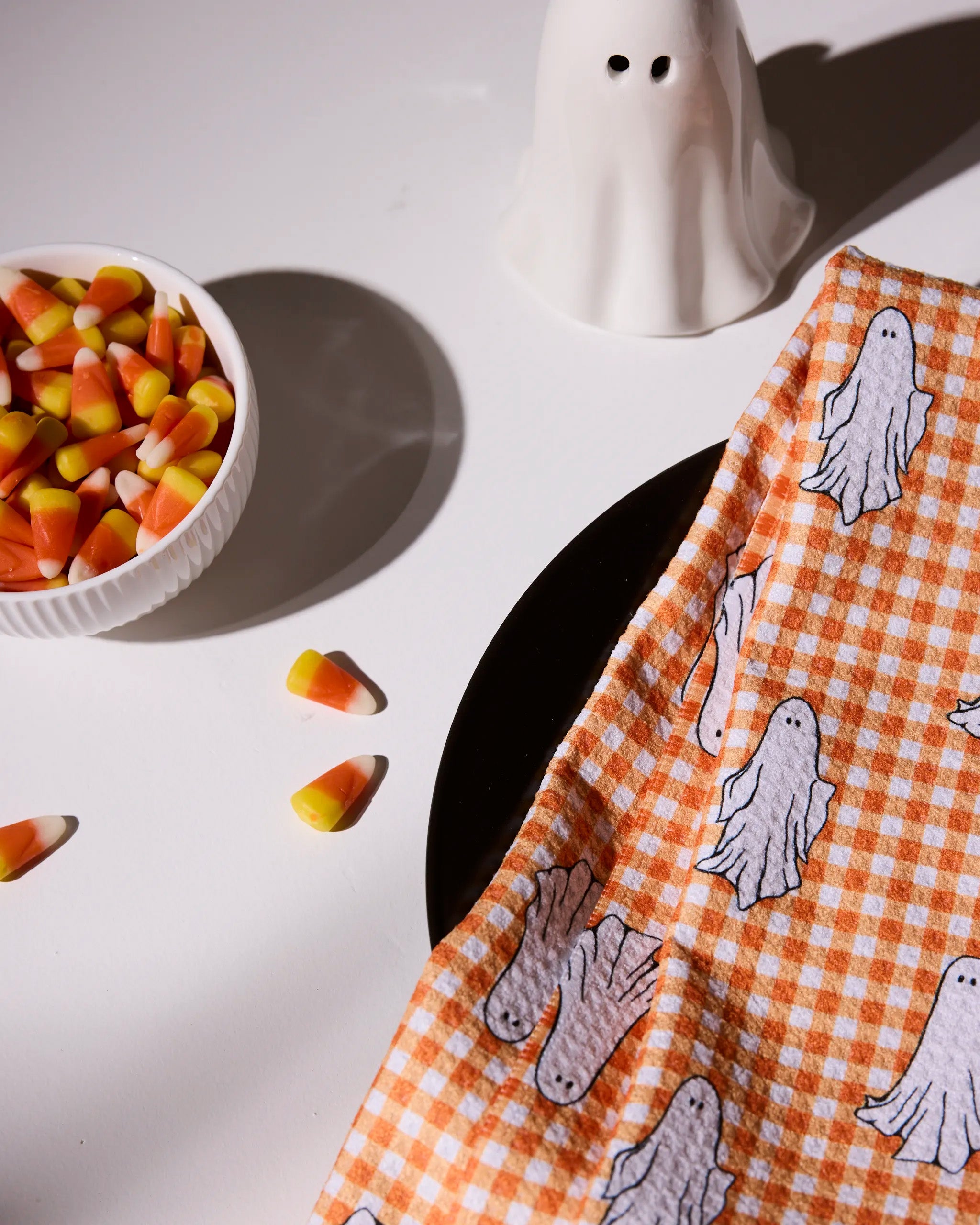 A bowl of candy corn, a ceramic ghost, and an orange checkered cloth with ghost prints are arranged on a white surface.