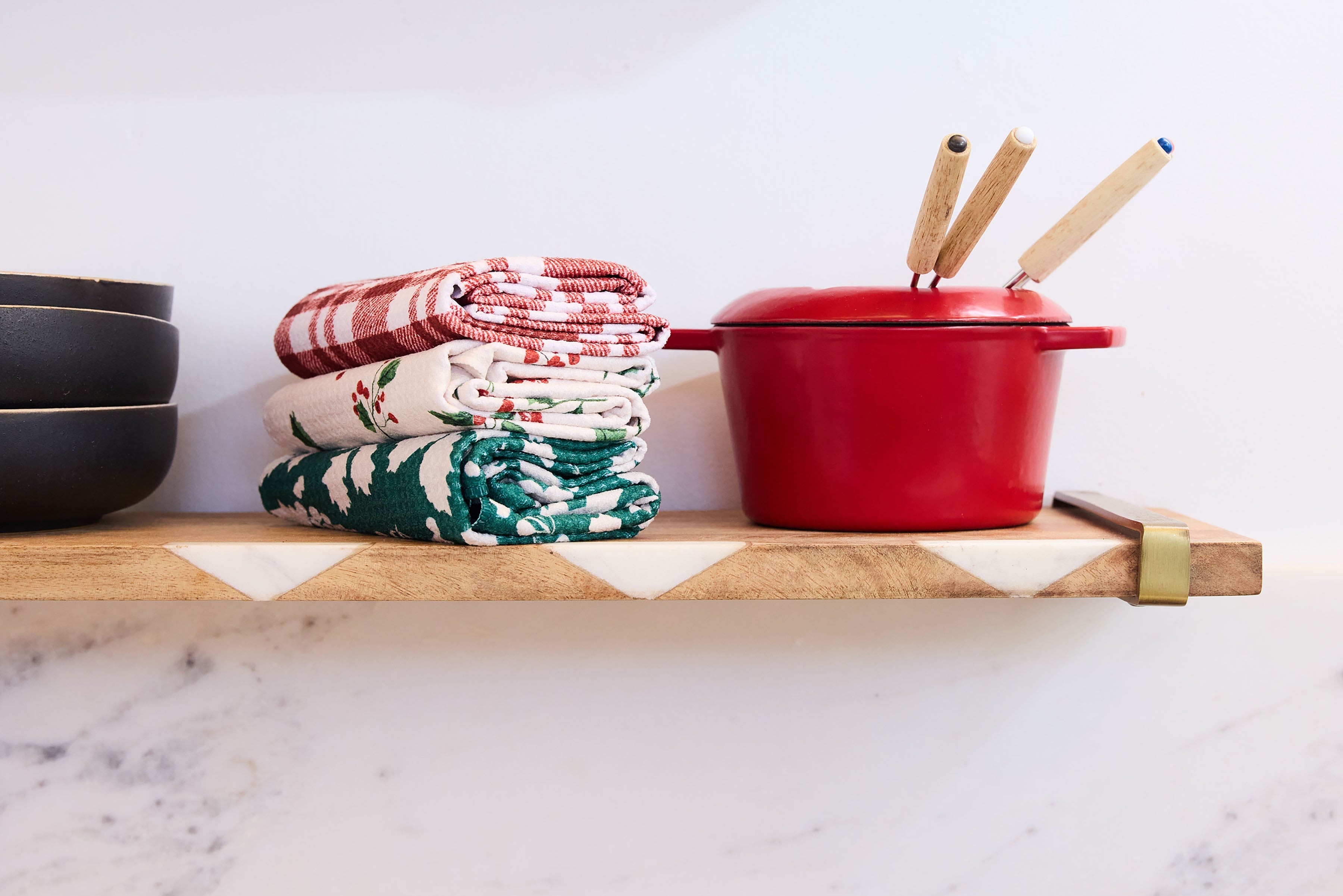 A wooden shelf holds stacked bowls, two piles of folded kitchen towels with red and green patterns, and a small red pot with two wooden-handled utensils inside.