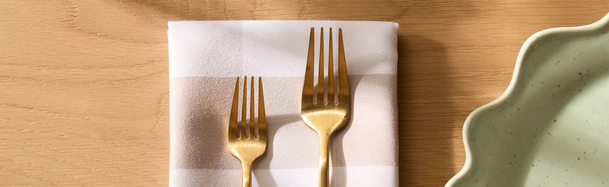 Two hands set a table with patterned napkins, gold cutlery, a black plate, a drink with a cinnamon stick, and small pinecones on a cream tablecloth.