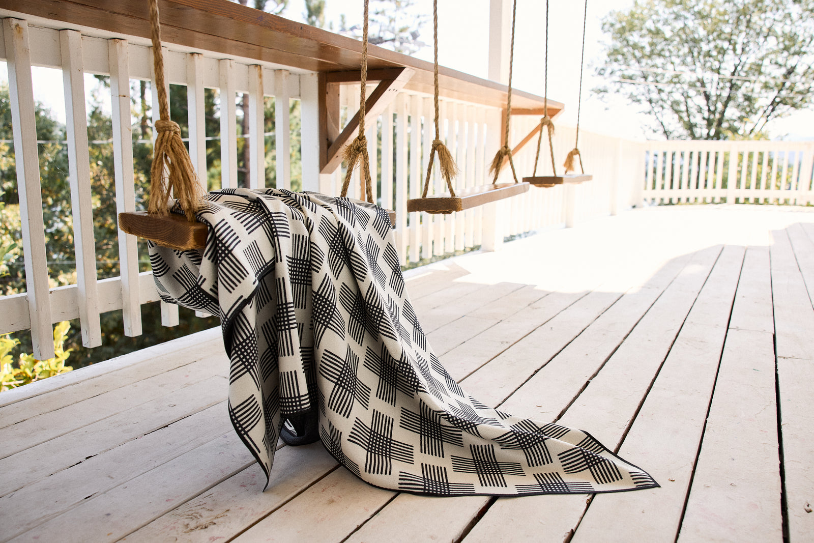 A black-and-white patterned blanket draped over a wooden swing on a spacious, sunlit porch with white railings and wooden floorboards. Trees are visible in the background.