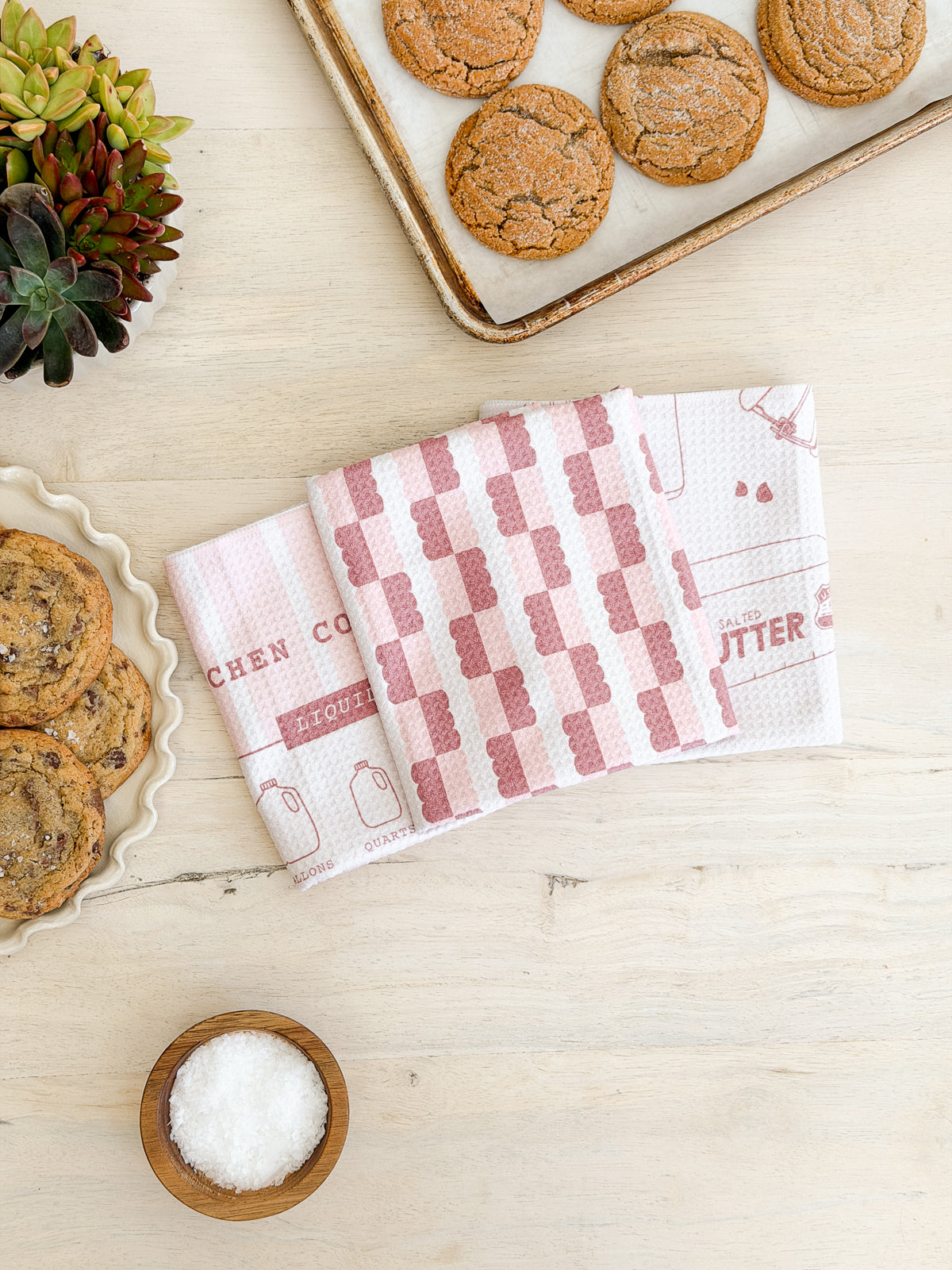 Three red-and-white dish towels are laid out on a light wooden table near a plate of cookies, a tray of cookies, a bowl of salt, and a potted succulent plant.
