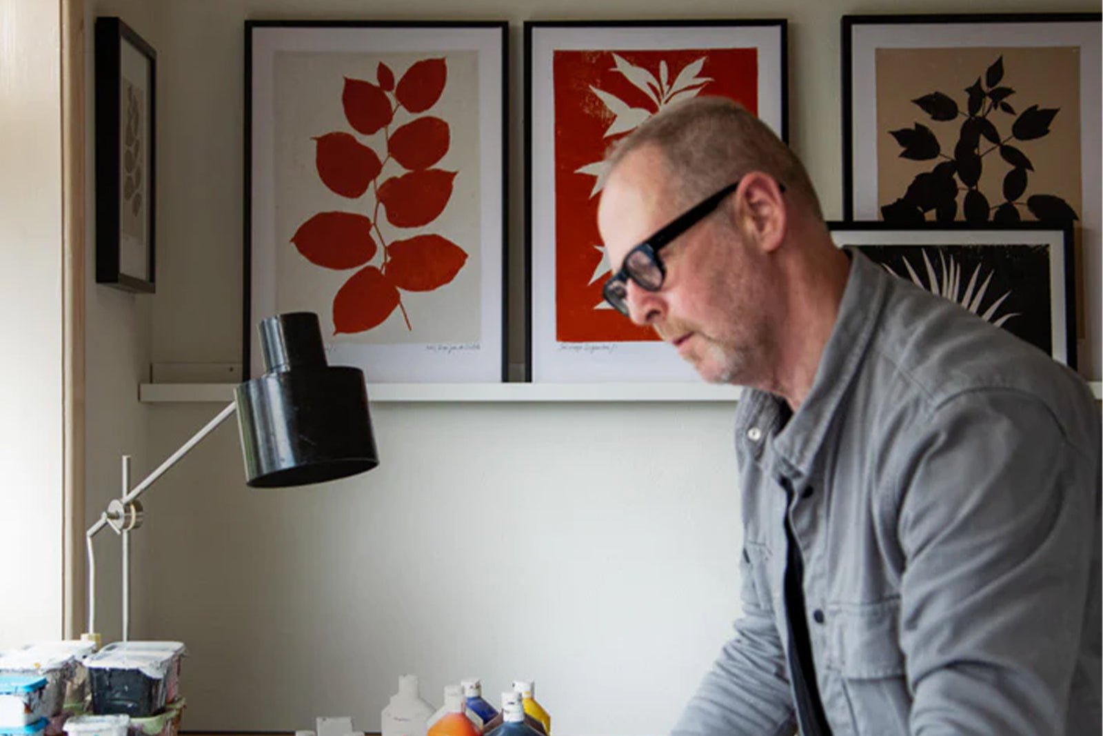 A man wearing glasses works at a desk in a room decorated with framed botanical prints featuring red and black leaves. A black desk lamp and art supplies are also visible on the desk.