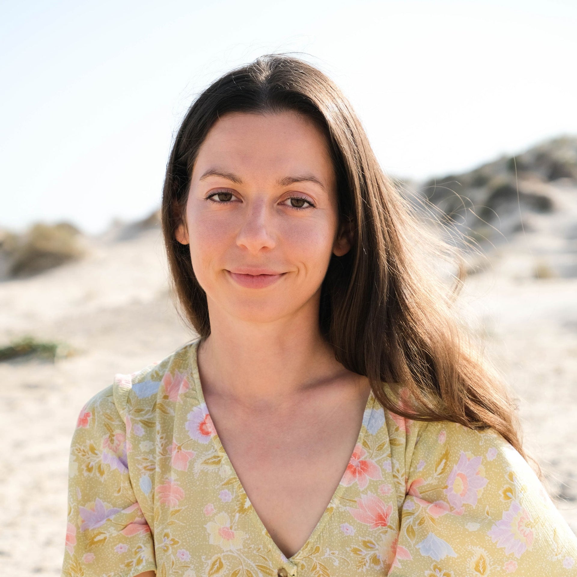 A woman with long brown hair wearing a yellow floral dress smiles gently while sitting outdoors on a sandy beach with dunes in the background.