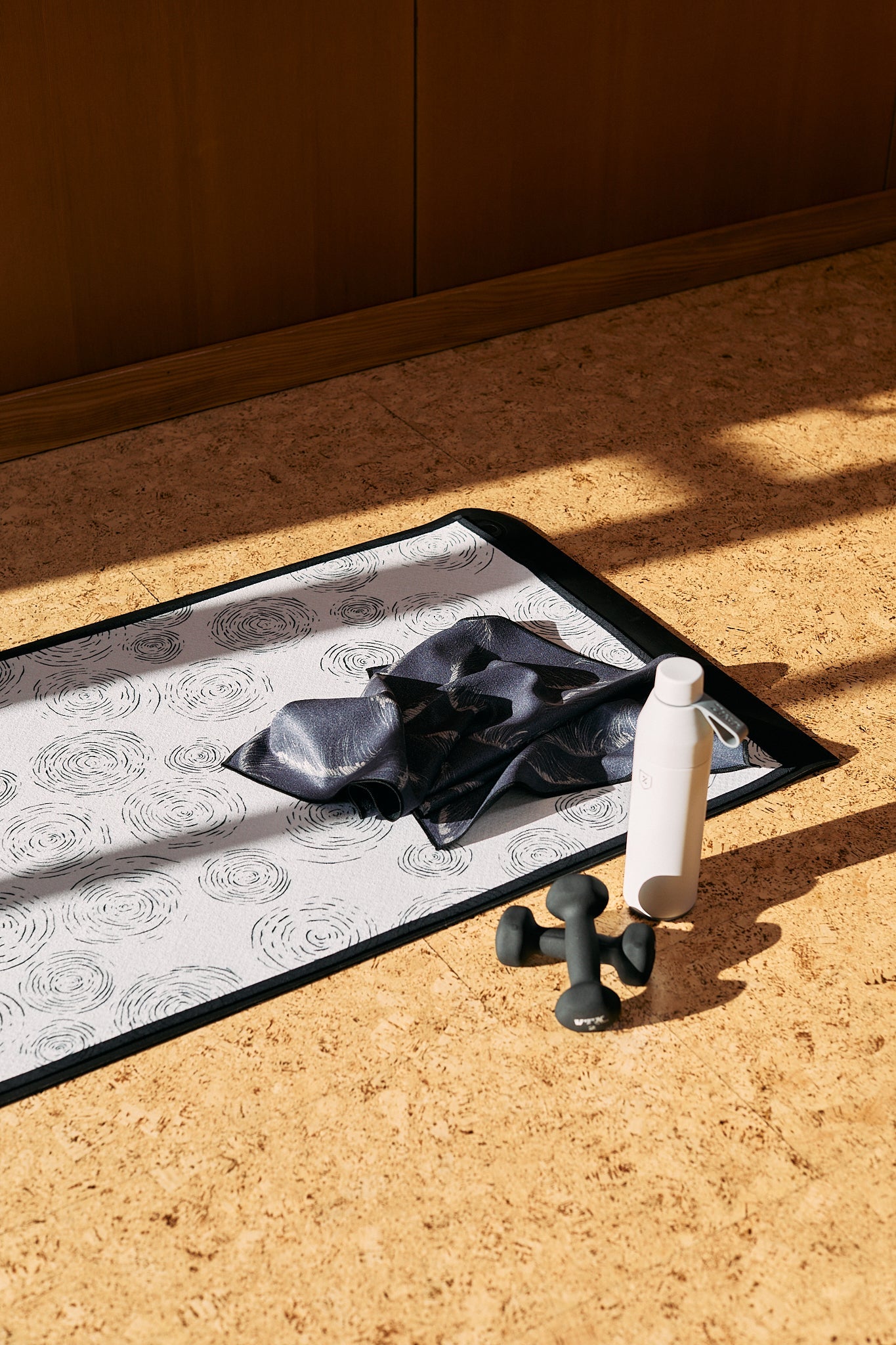 A patterned yoga mat on a cork floor with a black towel, a white water bottle, and a pair of small black dumbbells, lit by sunlight.