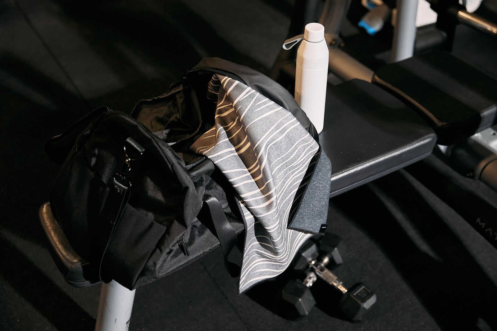 A gym bench holds a black bag, a striped towel, a white water bottle, and a pair of dumbbells, all set on a dark gym floor.