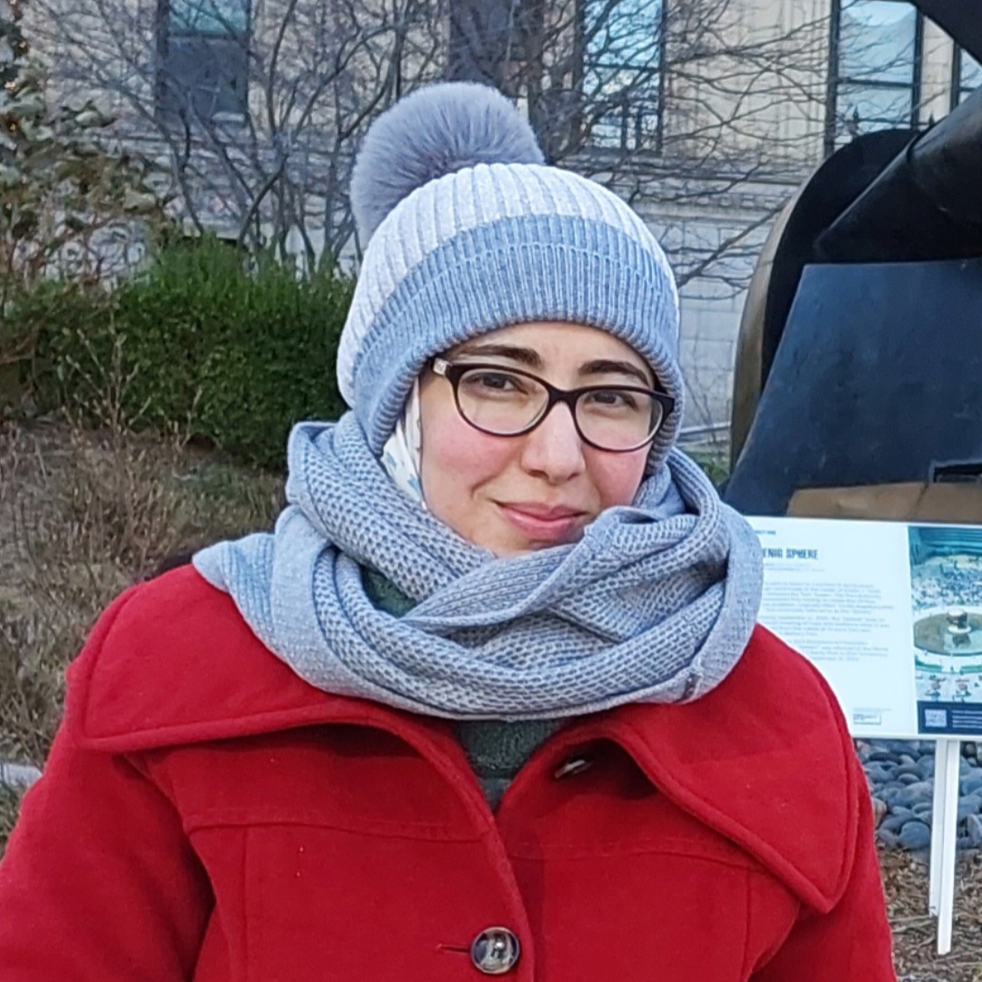 A person wearing glasses, a red coat, and a gray winter hat and scarf smiles outdoors in front of winter bushes and a sign.