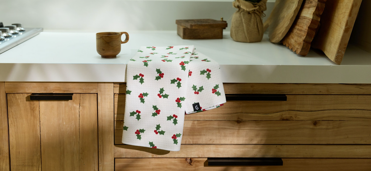 A white kitchen towel with a red and green holly berry pattern draped over a wooden drawer handle on a kitchen counter.