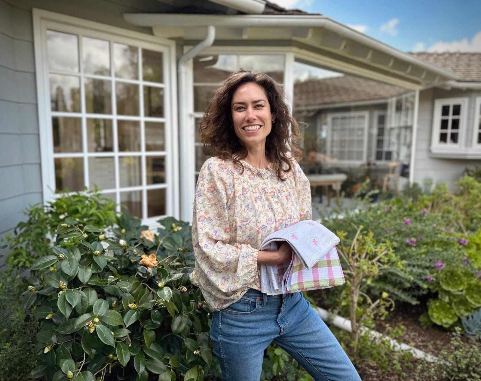 A woman with curly hair stands outside a house, smiling and holding folded towels. She wears a floral blouse and jeans, surrounded by green plants and flowers in a garden.
