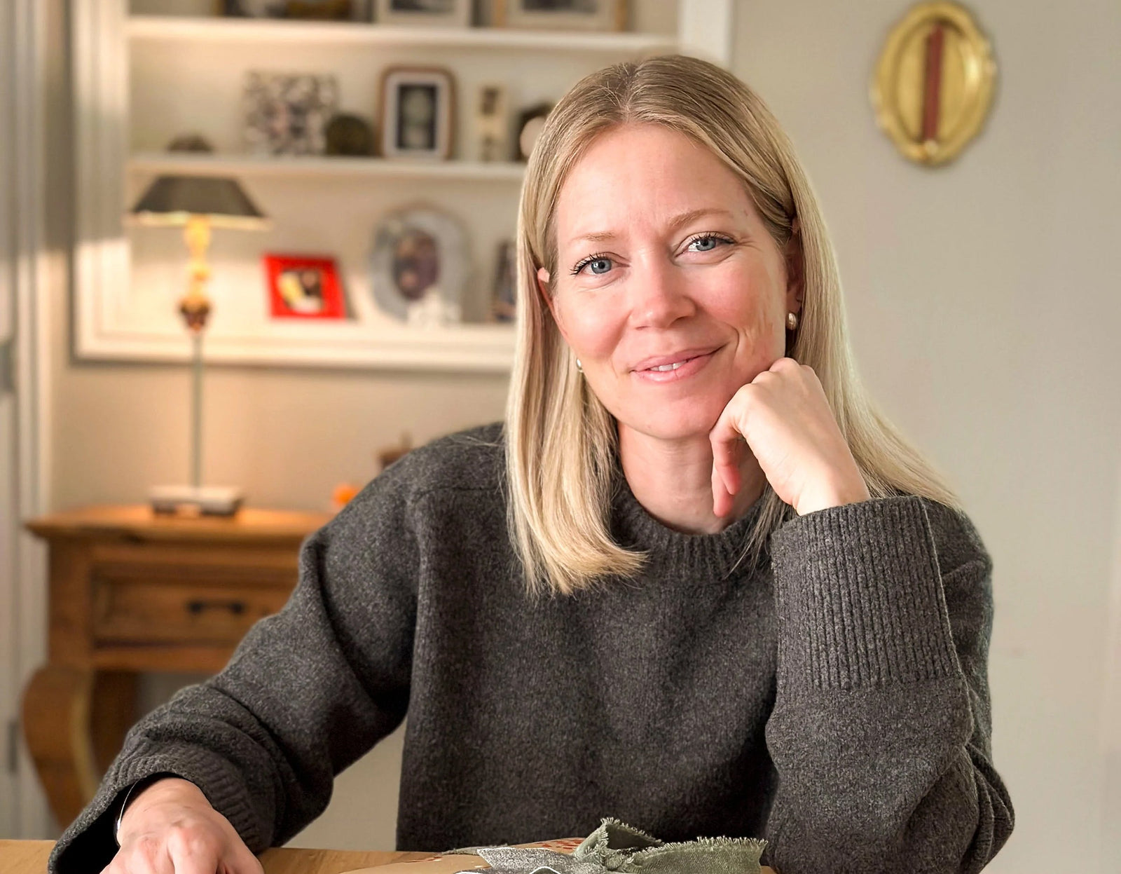 A woman with blonde hair wearing a dark sweater sits at a table, smiling gently with her hand resting on her chin. A wooden shelf and framed photos are visible in the background.