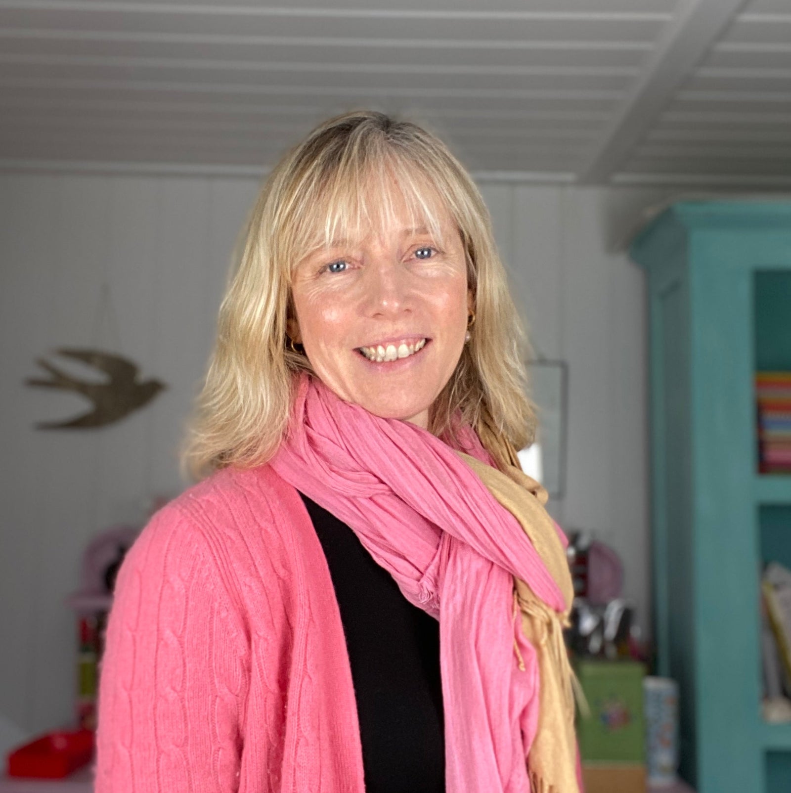 A woman with blonde hair wearing a pink scarf and sweater smiles at the camera in a cozy, well-lit room with a teal cabinet and a bird decoration on the wall.