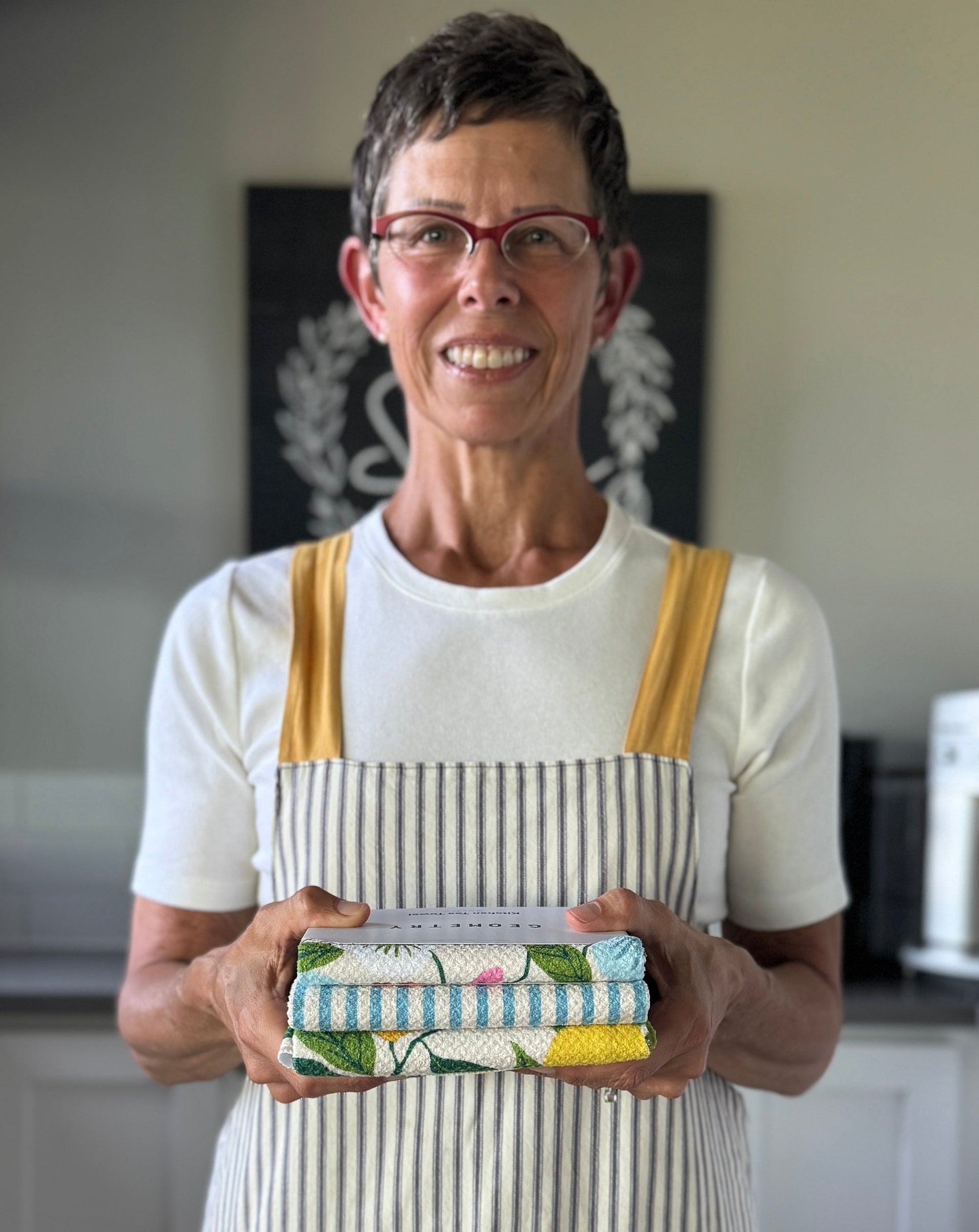 A smiling person wearing glasses, a white shirt, and a striped apron stands in a kitchen holding a stack of colorful folded dish towels.
