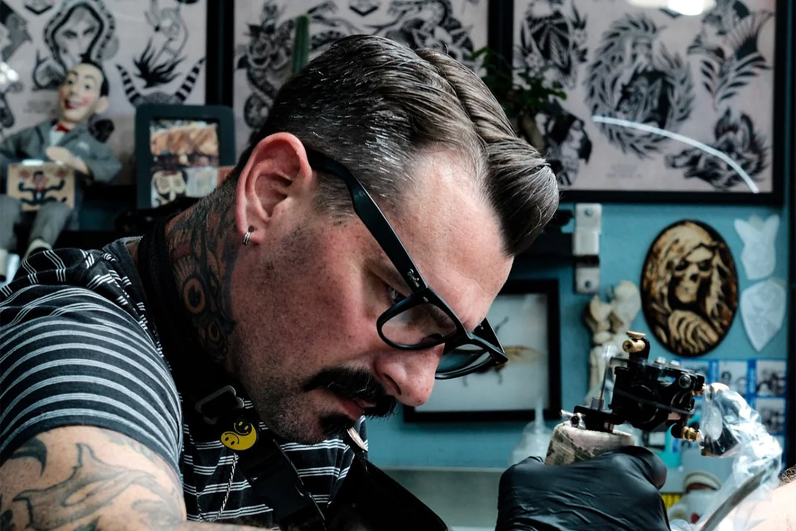 A tattoo artist with glasses and tattoos works intently on a client, holding a tattoo machine. The background displays various tattoo designs and artwork on the wall.