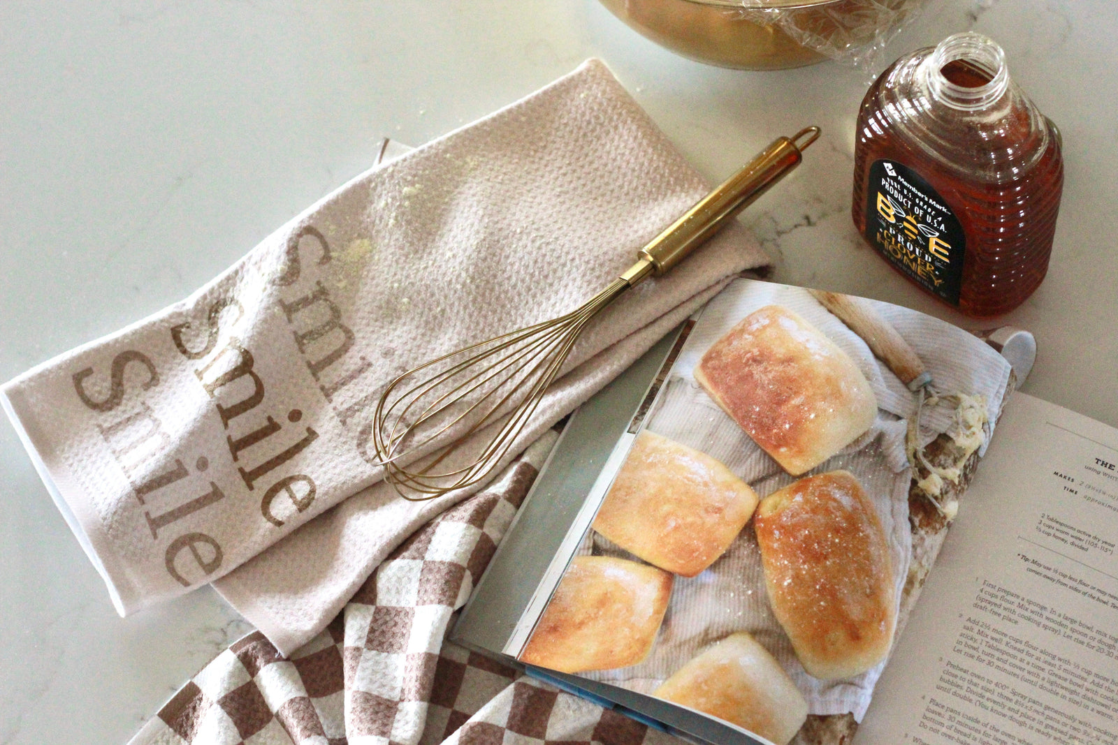 A whisk, honey bottle, two kitchen towels, and an open cookbook displaying bread rolls are arranged on a light countertop.