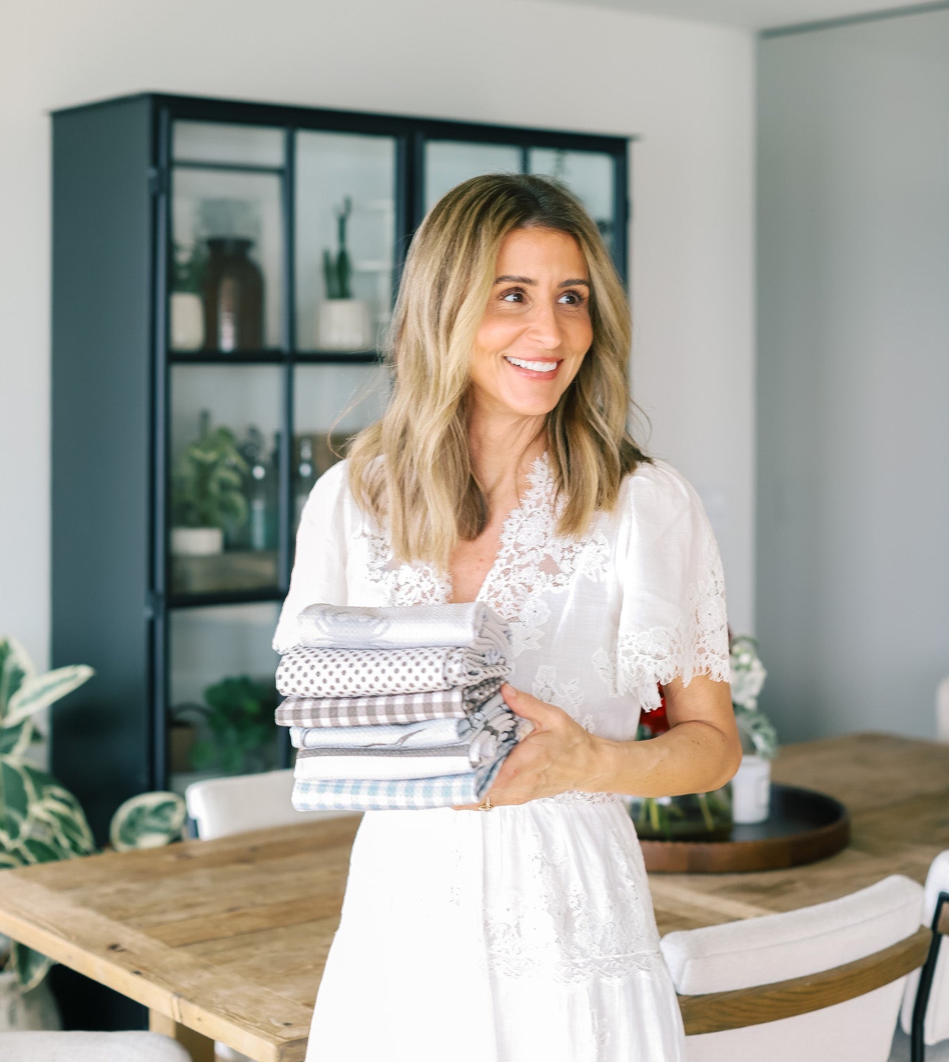 A smiling woman in a white dress stands indoors, holding a stack of folded kitchen towels. She is in a bright, modern dining room with wooden furniture and plants in the background.
