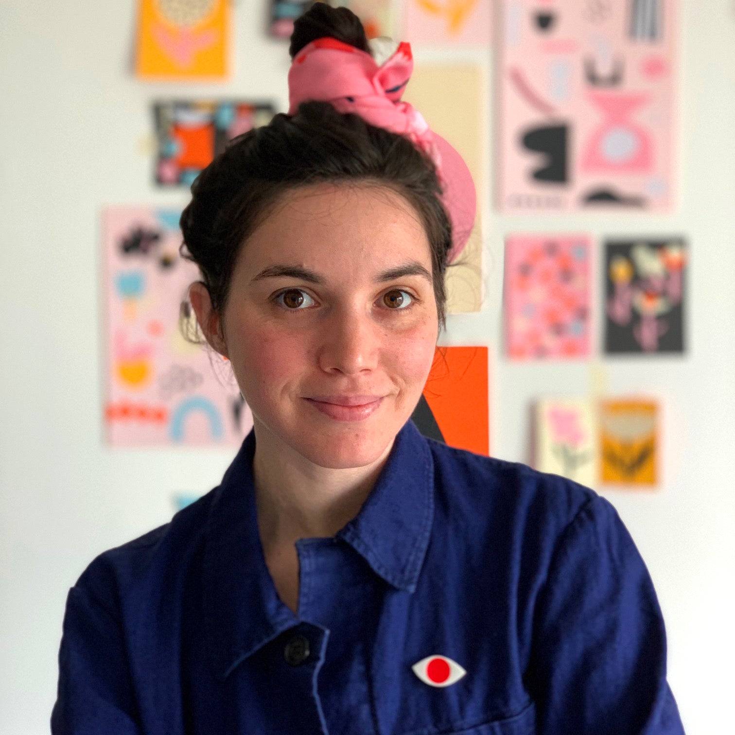 A woman with dark hair in a pink headband and blue jacket smiles softly; colorful art posters are blurred in the background.