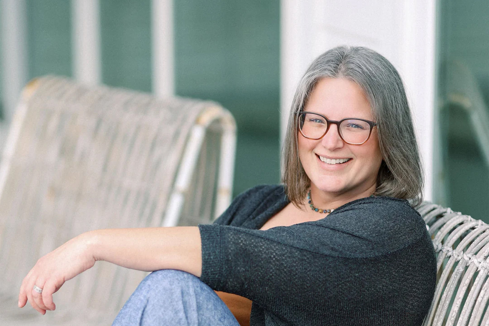 A smiling woman with gray hair and glasses sits on a wicker chair, wearing a dark top and light blue pants, with her arm resting on the chair’s back.