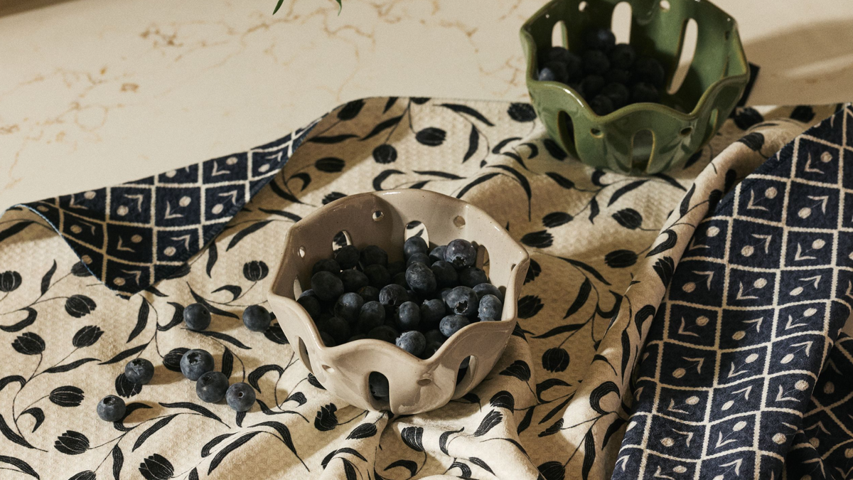Two ceramic berry baskets—one beige, one green—filled with blueberries sit on a patterned blue and white fabric on a light-colored countertop. Some loose blueberries are scattered around.