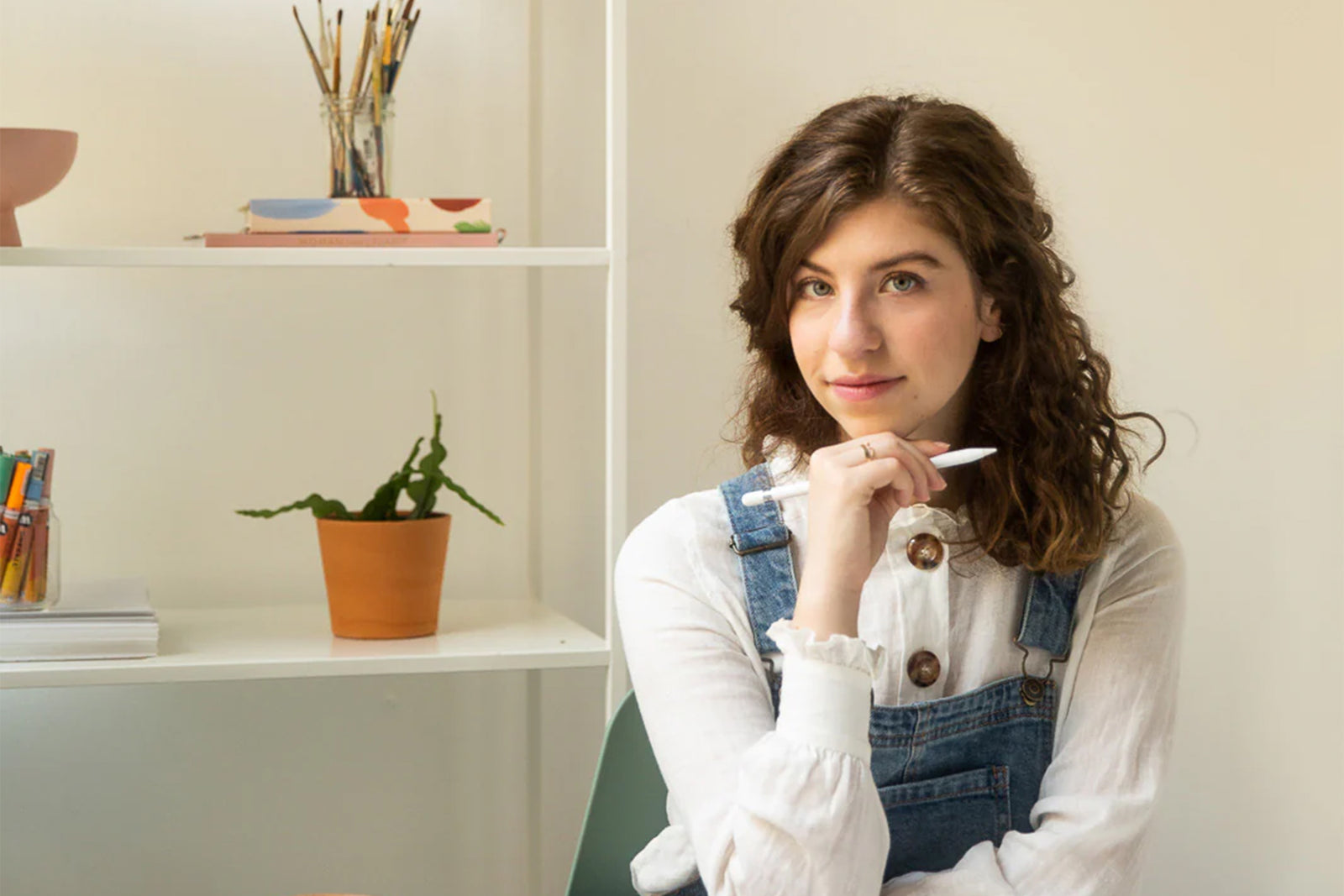 A woman with curly brown hair wearing a white shirt and denim overalls sits indoors, holding a pen and smiling slightly. Shelves with books, art supplies, and a potted plant are in the background.