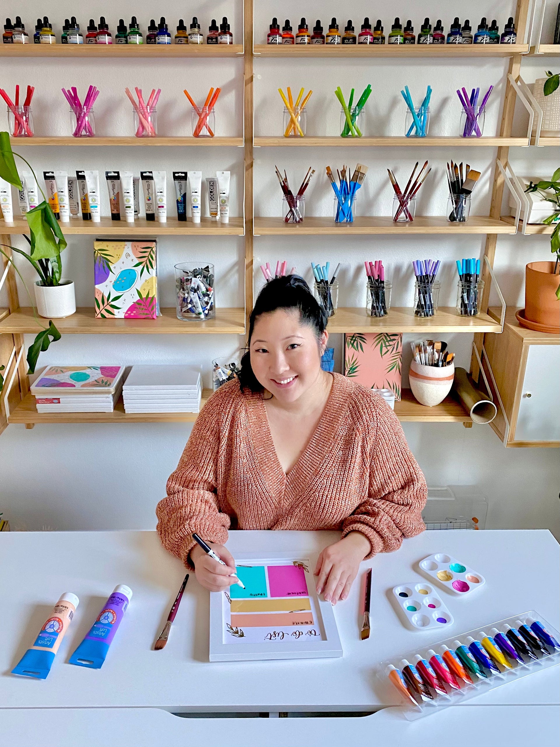 A woman smiles while drawing at a white desk, surrounded by art supplies such as paints, markers, and brushes, with colorful shelves of art materials behind her.