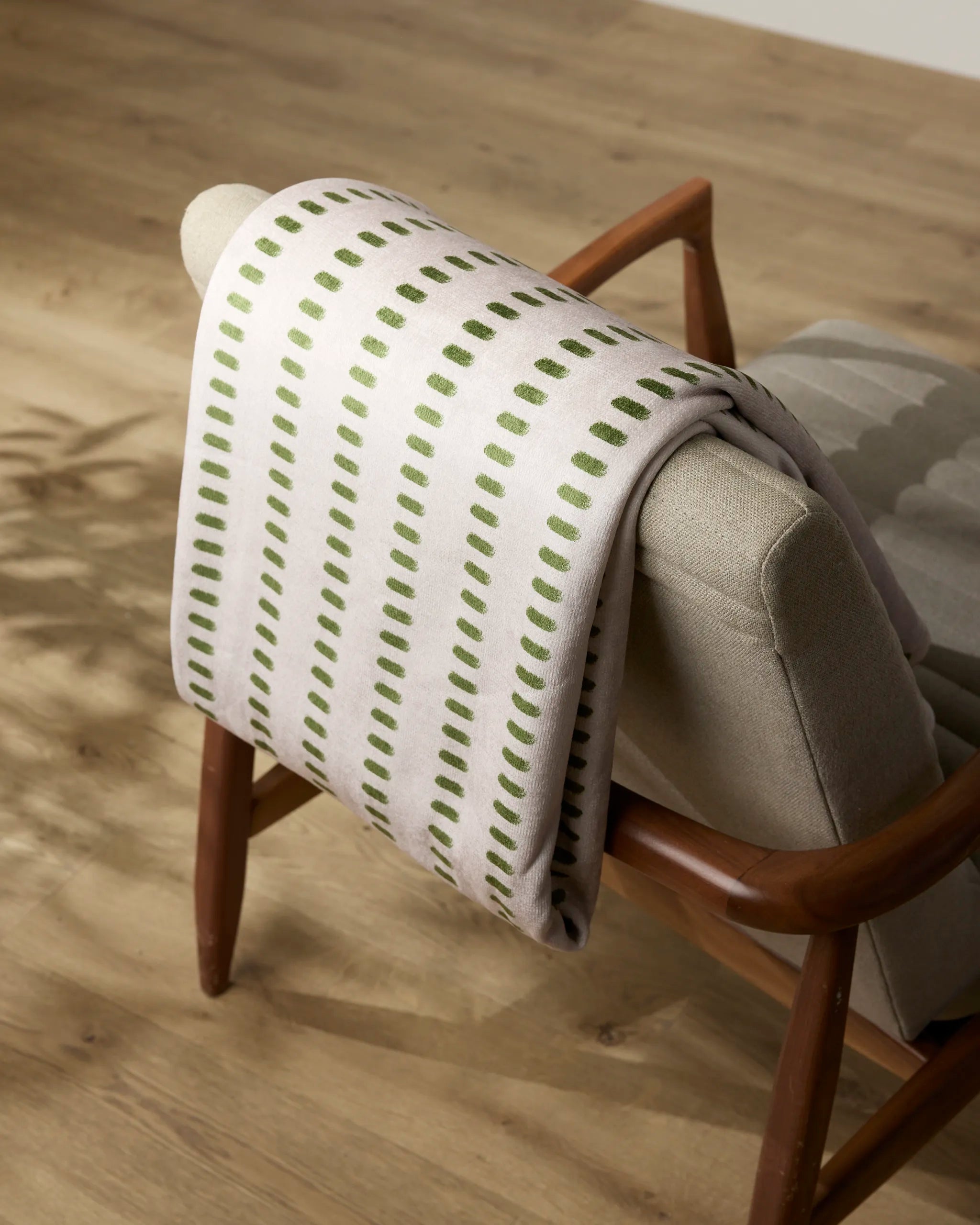 A beige chair with wooden arms has a folded white blanket draped over the backrest. The blanket features a pattern of green, evenly spaced vertical dashes.