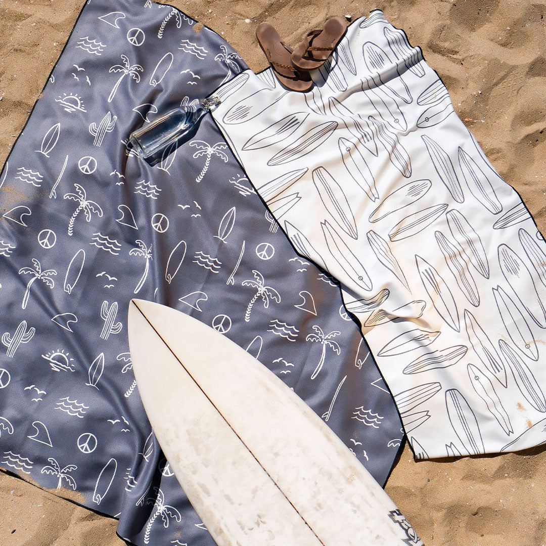 Two patterned towels, one gray with palm trees and peace signs, and one white with surfboards, are spread on sand. A surfboard and sandals are placed nearby.