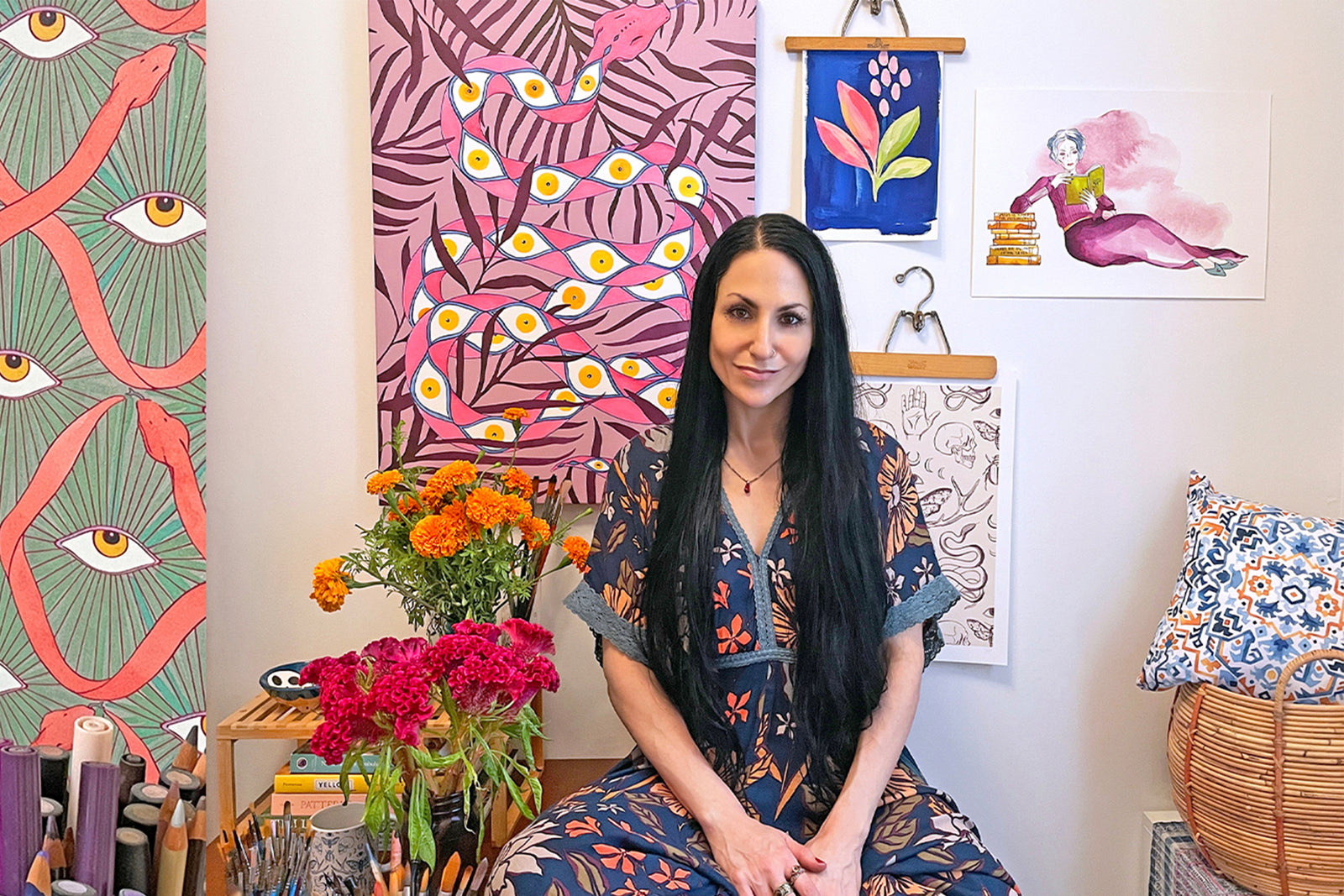 A woman with long dark hair sits smiling in a colorful art studio, surrounded by vibrant paintings, flowers, art supplies, and patterned decor.