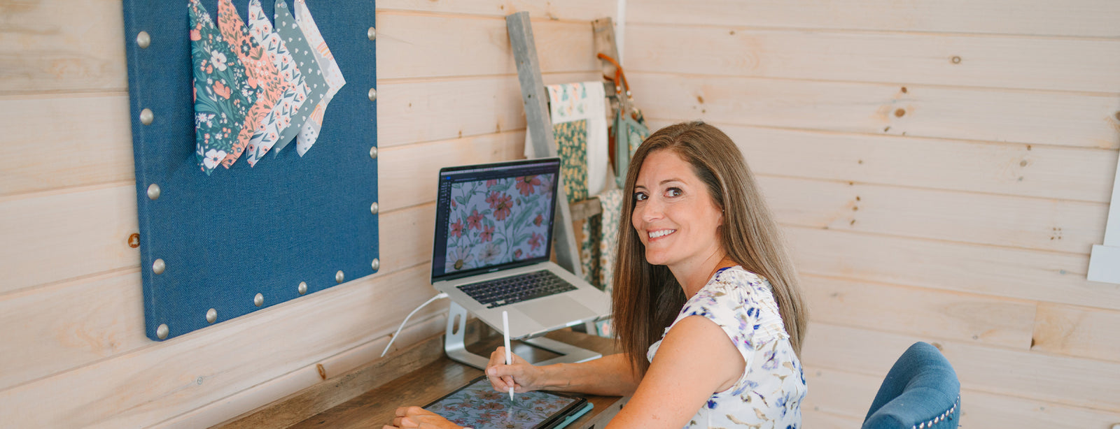 A woman sits at a wooden desk using a digital tablet and stylus, smiling at the camera. A laptop displaying floral designs and fabric swatches are visible in her workspace.
