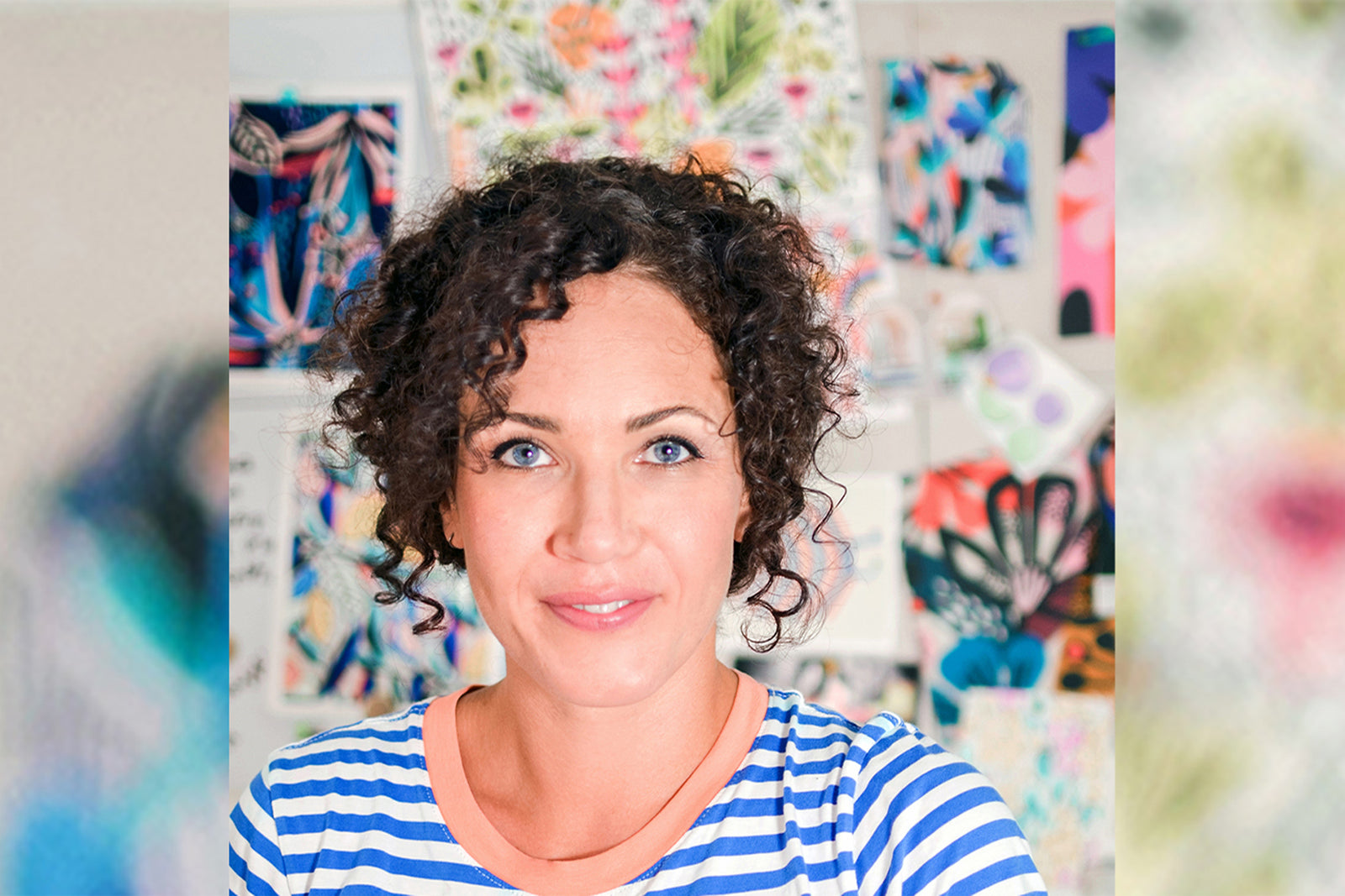 A woman with curly brown hair and blue eyes is smiling, wearing a blue-and-white striped shirt. She stands in front of a wall covered with colorful, abstract art pieces.