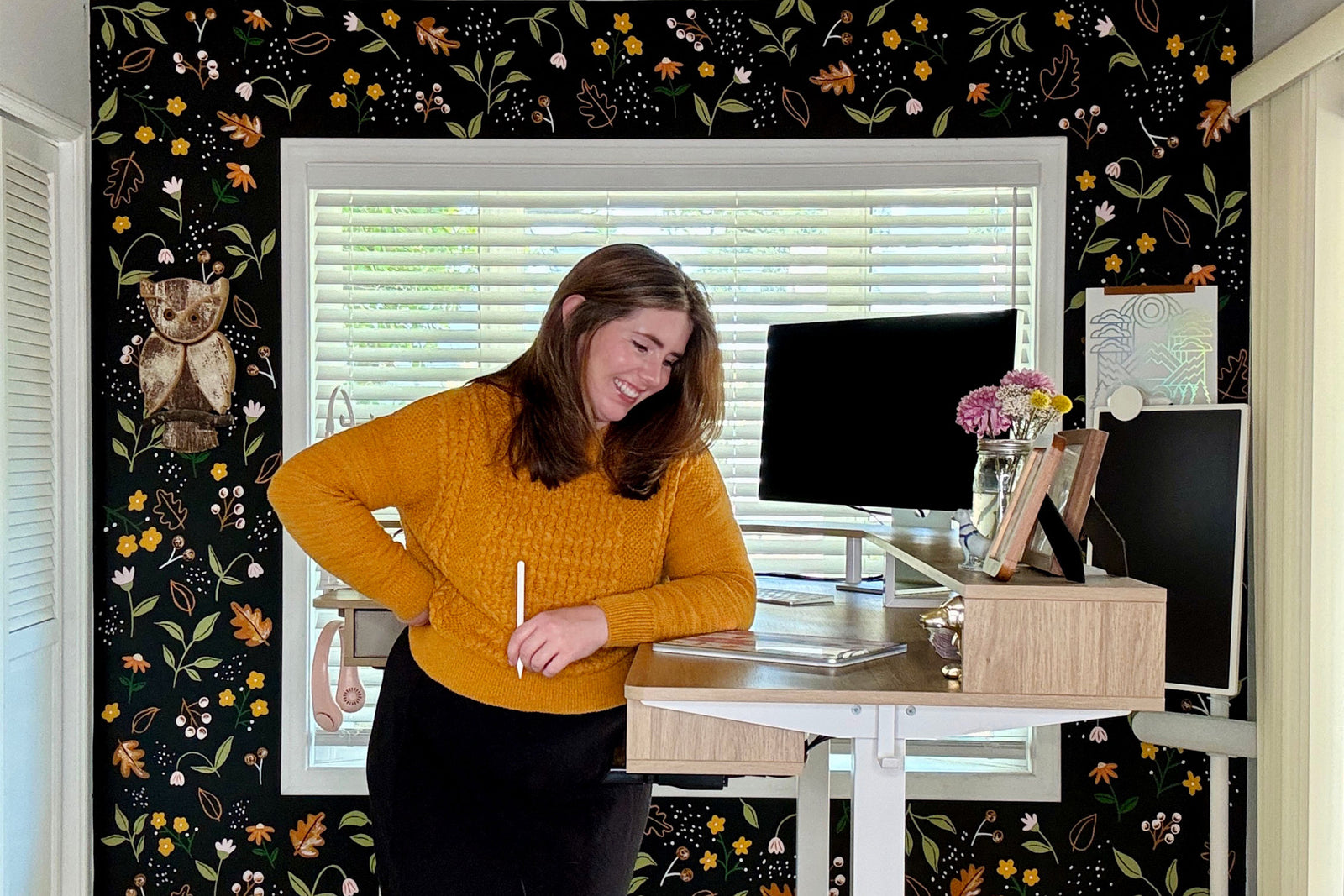 A woman in a mustard yellow sweater stands and smiles at a standing desk with a computer, flowers, and a tablet in a home office with floral wallpaper and an owl decoration.