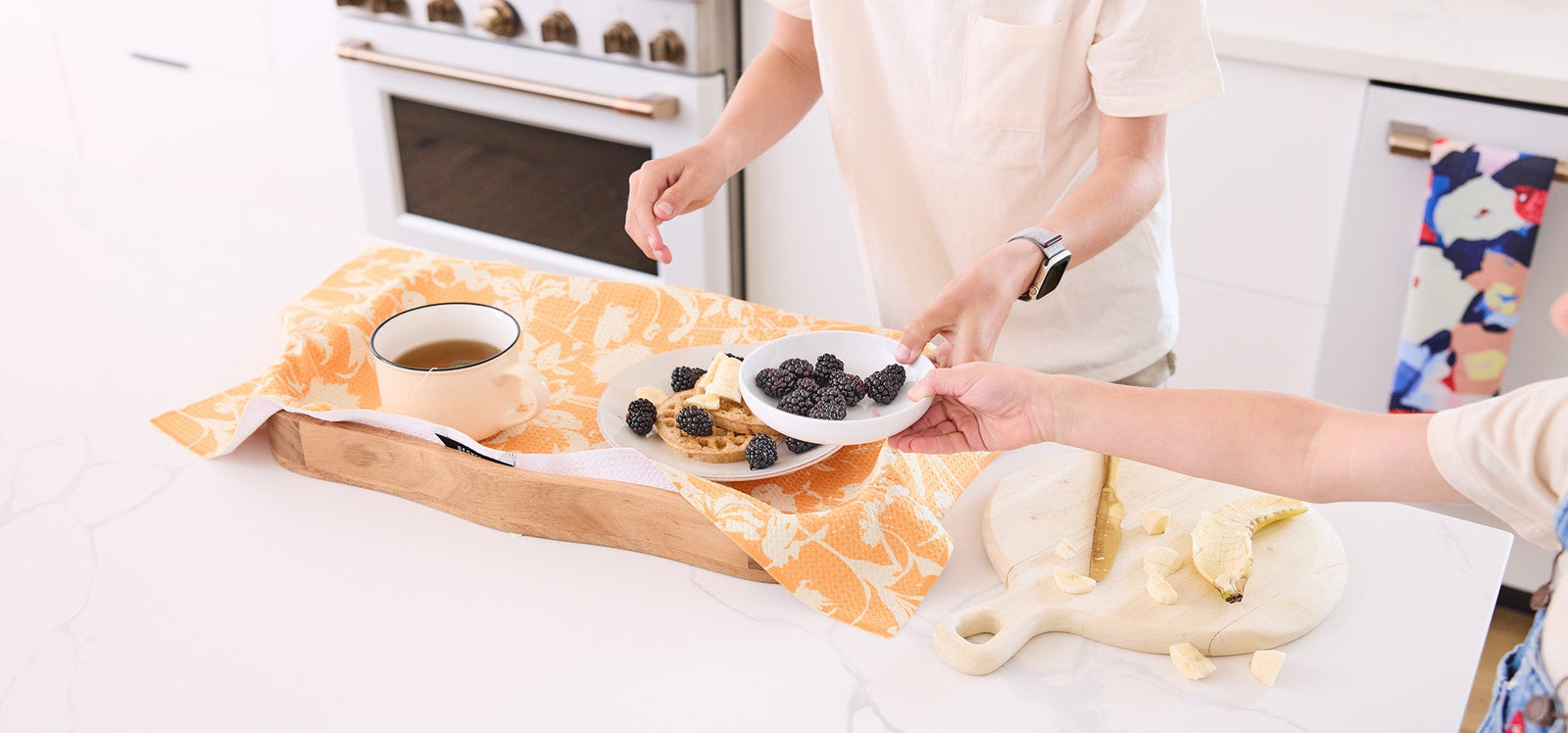 Two people share a bowl of blackberries over a white kitchen counter with a wooden tray, cup of tea, cookies, a yellow cloth, and a cutting board with banana slices.