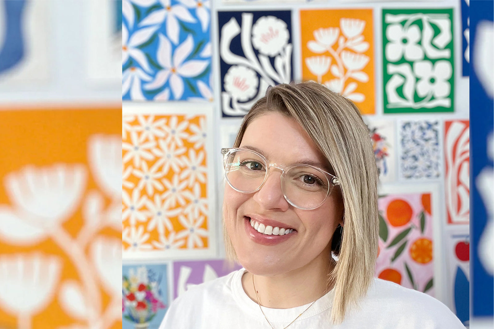 A smiling person with glasses and blond hair stands in front of a colorful wall covered with floral and abstract art in orange, blue, green, and white tones.