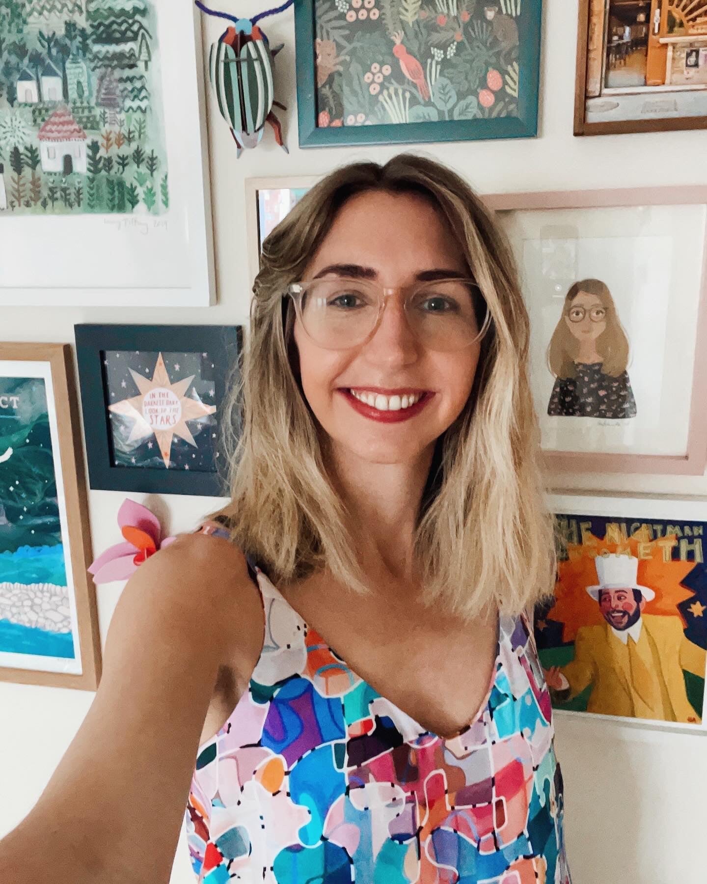 A smiling woman with blonde hair and glasses takes a selfie in front of a wall decorated with colorful framed artwork and illustrations. She is wearing a patterned, multicolored top.