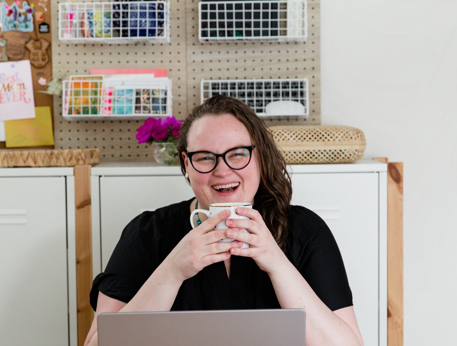 A person with glasses and long hair smiles while holding a mug, sitting at a desk with a laptop. Behind them are shelves with colorful supplies and a bouquet of purple flowers.