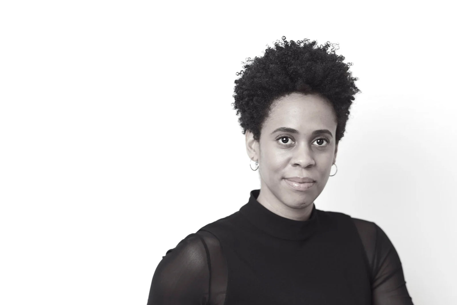 A woman with short curly hair and hoop earrings, wearing a black top with sheer sleeves, stands against a plain white background, looking into the camera with a slight smile.