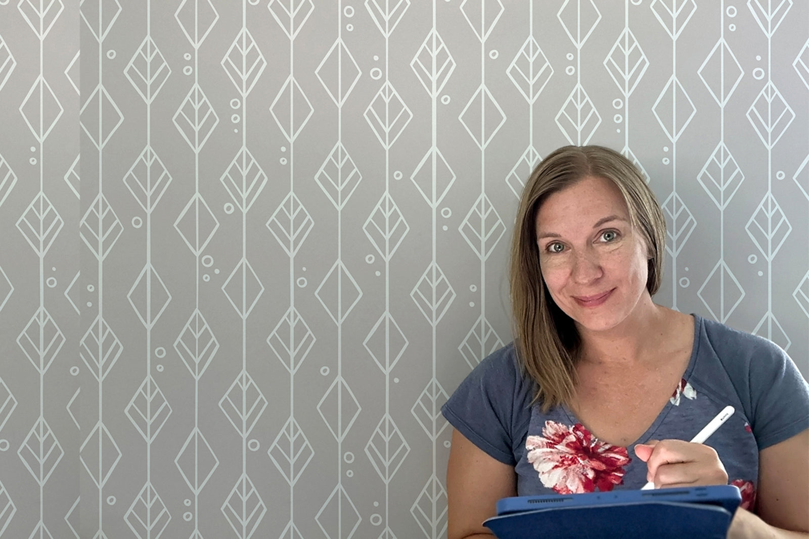 A woman with straight, shoulder-length hair smiles while writing on a clipboard. She wears a blue top with a floral pattern, standing in front of a gray wall with a geometric leaf design.