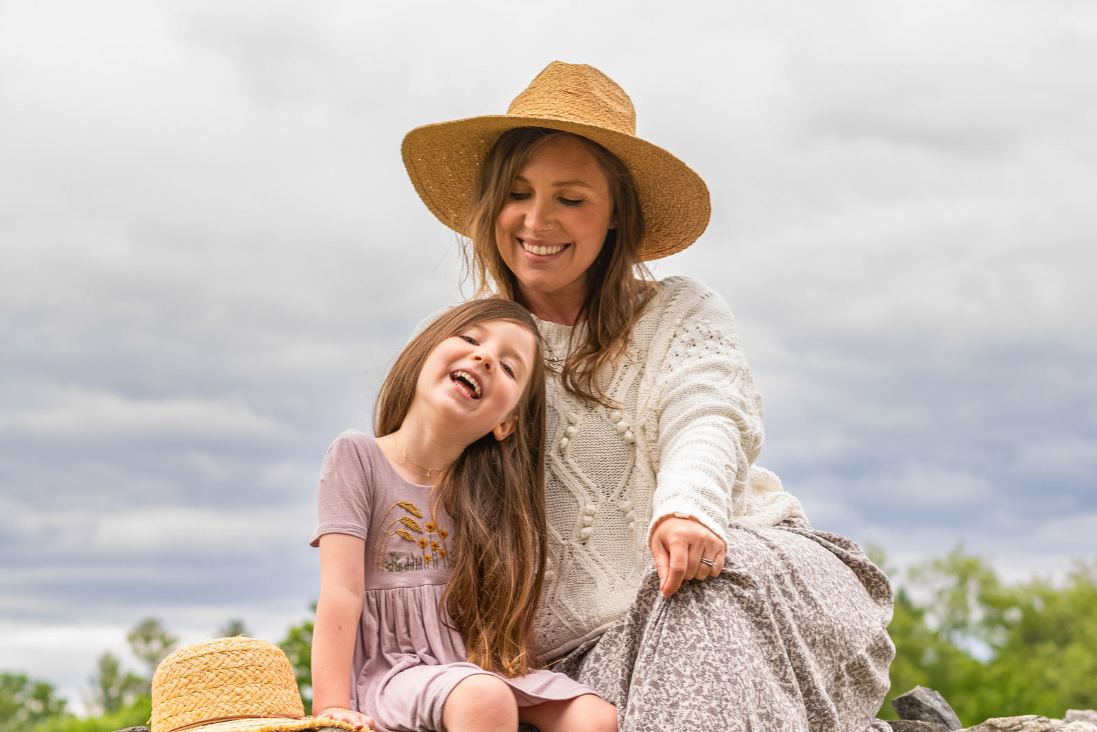 A woman in a straw hat sits outdoors with a young girl, both smiling and enjoying a cloudy day. The girl leans against the woman, and a second hat rests nearby on the grass.