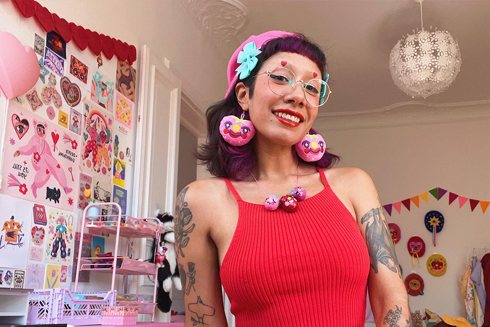 A smiling person with pink glasses, large pink donut earrings, and a red dress stands in a colorful, decorated room with wall art, crafts, and playful decor.
