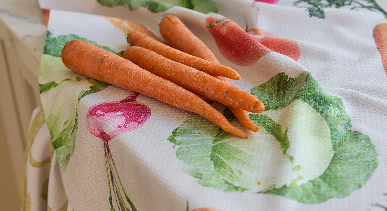 Several fresh carrots are lying on a colorful kitchen towel that is decorated with illustrations of radishes, lettuce, and other vegetables.