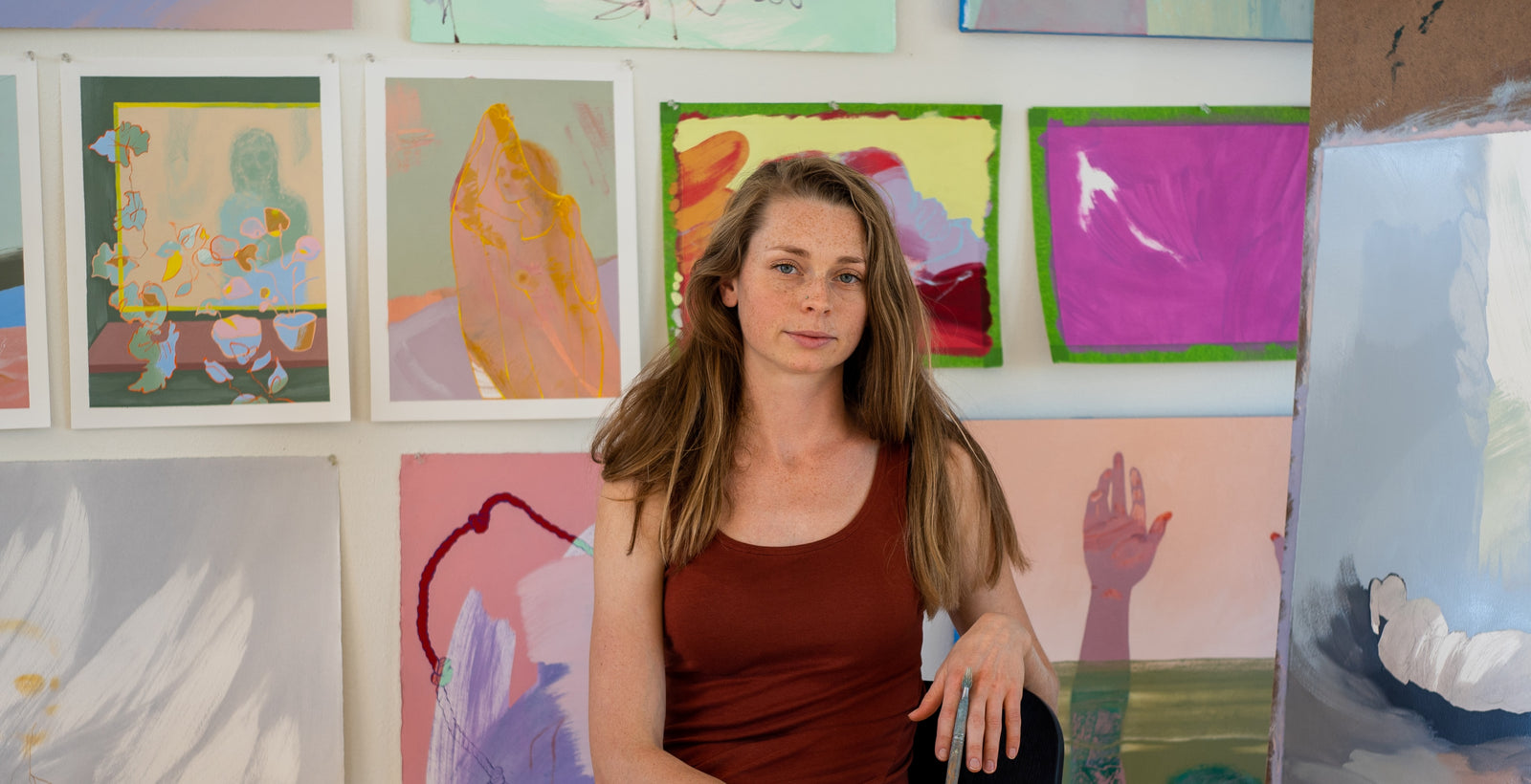 A woman with long brown hair and a rust-colored tank top sits in front of a wall displaying colorful abstract paintings and artworks.