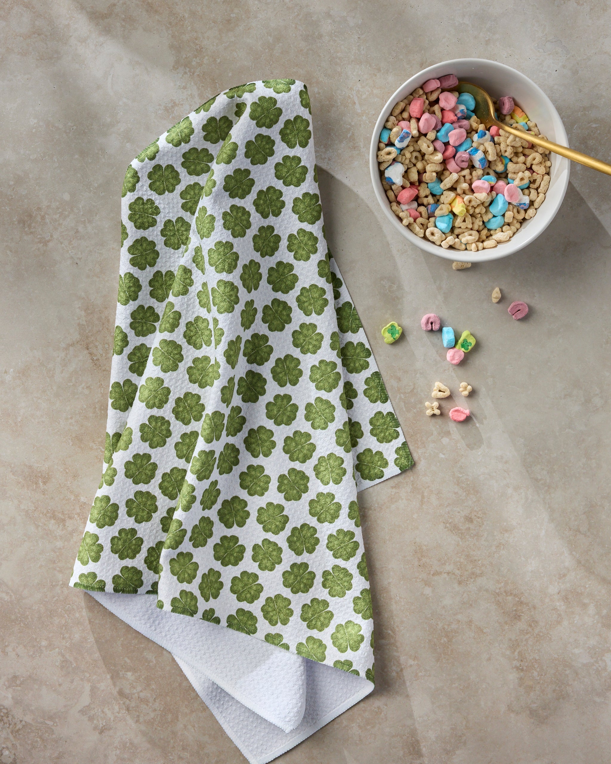 A white towel with green shamrock patterns lies next to a bowl of cereal with colorful marshmallows and a gold spoon. Several marshmallows are scattered on the light countertop.