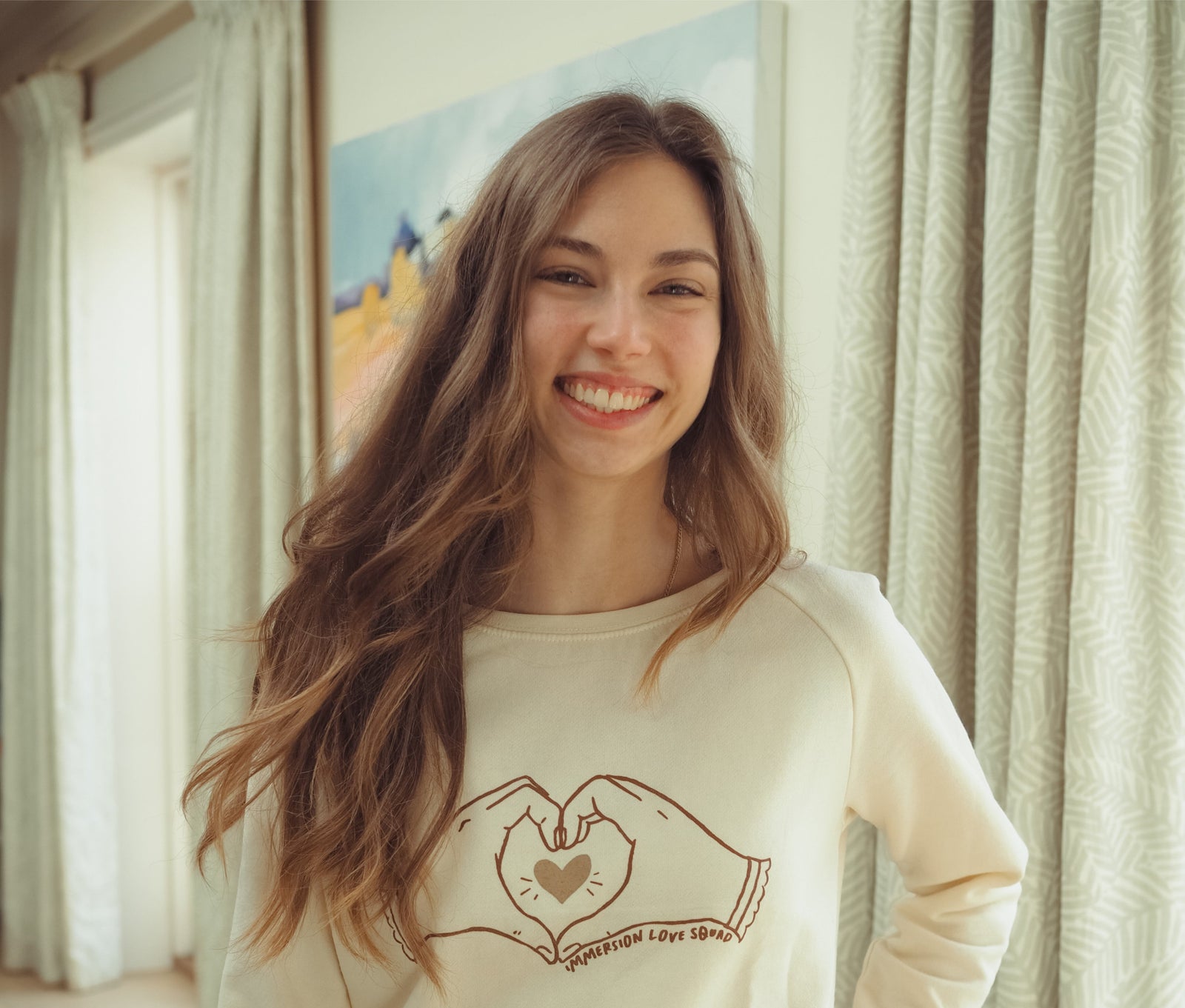 A young woman with long brown hair smiles indoors, wearing a cream-colored sweater with a drawing of hands forming a heart and the words Expression Love Squad. Light curtains and a painting are in the background.