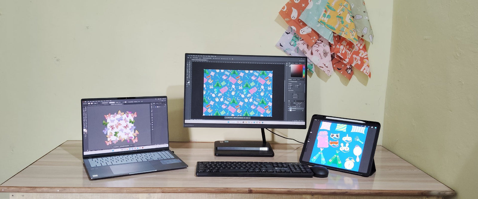 A workspace with a laptop, monitor, and tablet displaying colorful graphic designs on a wooden desk. Fabric swatches with similar patterns hang on a light yellow wall in the background.