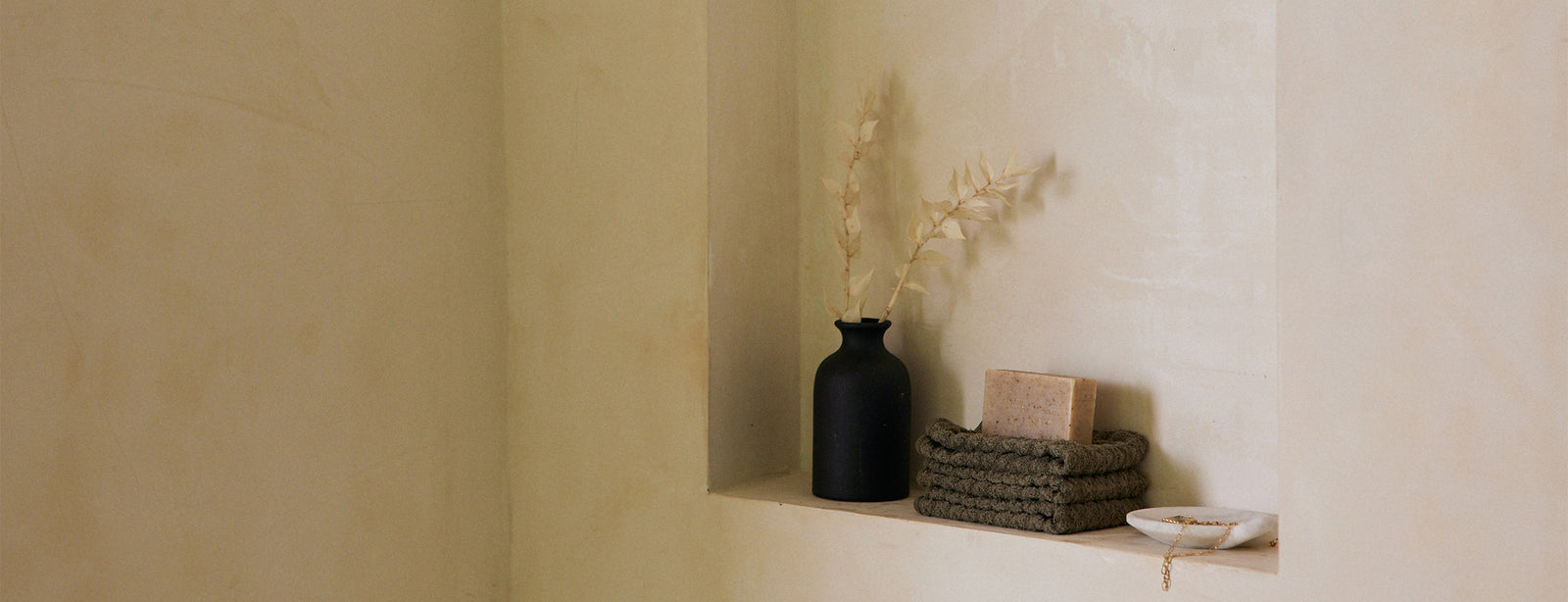 A minimalist beige wall niche holds a black vase with dried grasses, a coiled green mat, a rectangular soap, and a small white dish with a beaded bracelet.