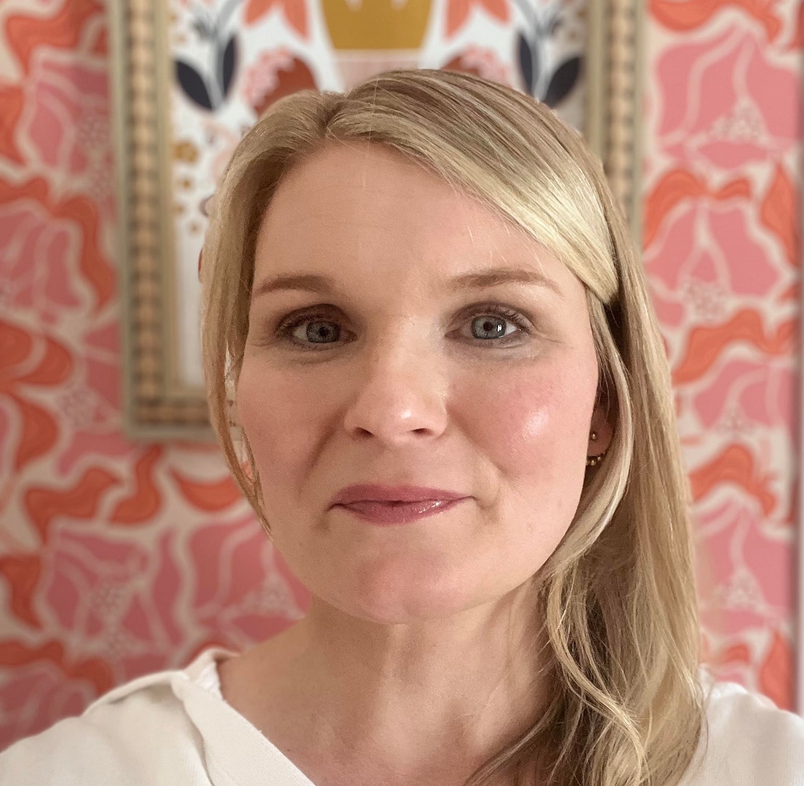 Blonde woman with blue eyes and pink lipstick smiles at the camera, against a backdrop of floral wallpaper and a framed artwork.