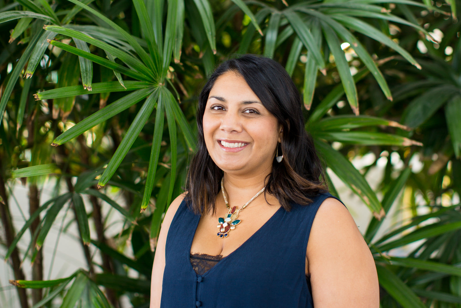 A woman with medium-length dark hair, wearing a navy sleeveless top and a statement necklace, smiles while standing in front of lush green tropical plants.