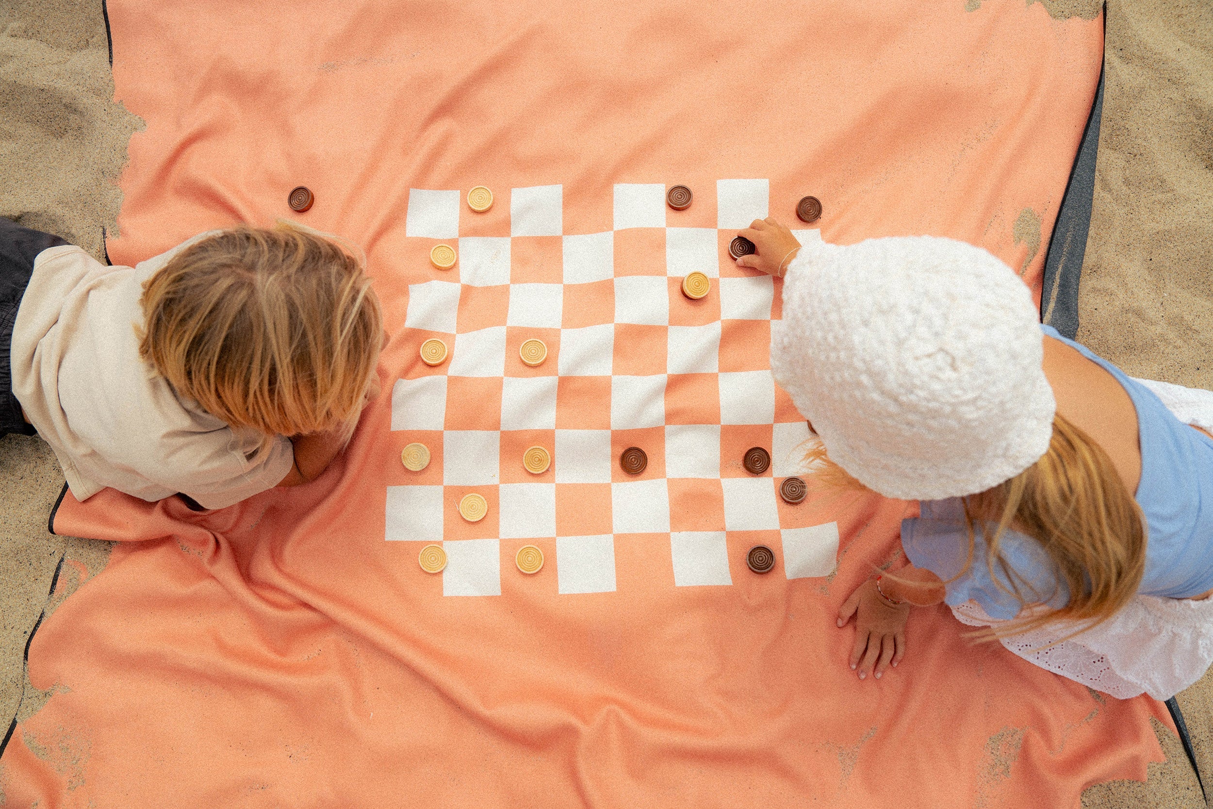 A split image: on the left, a checkerboard with limes and tangerines as pieces; on the right, three children play a board game on a blanket at the beach.