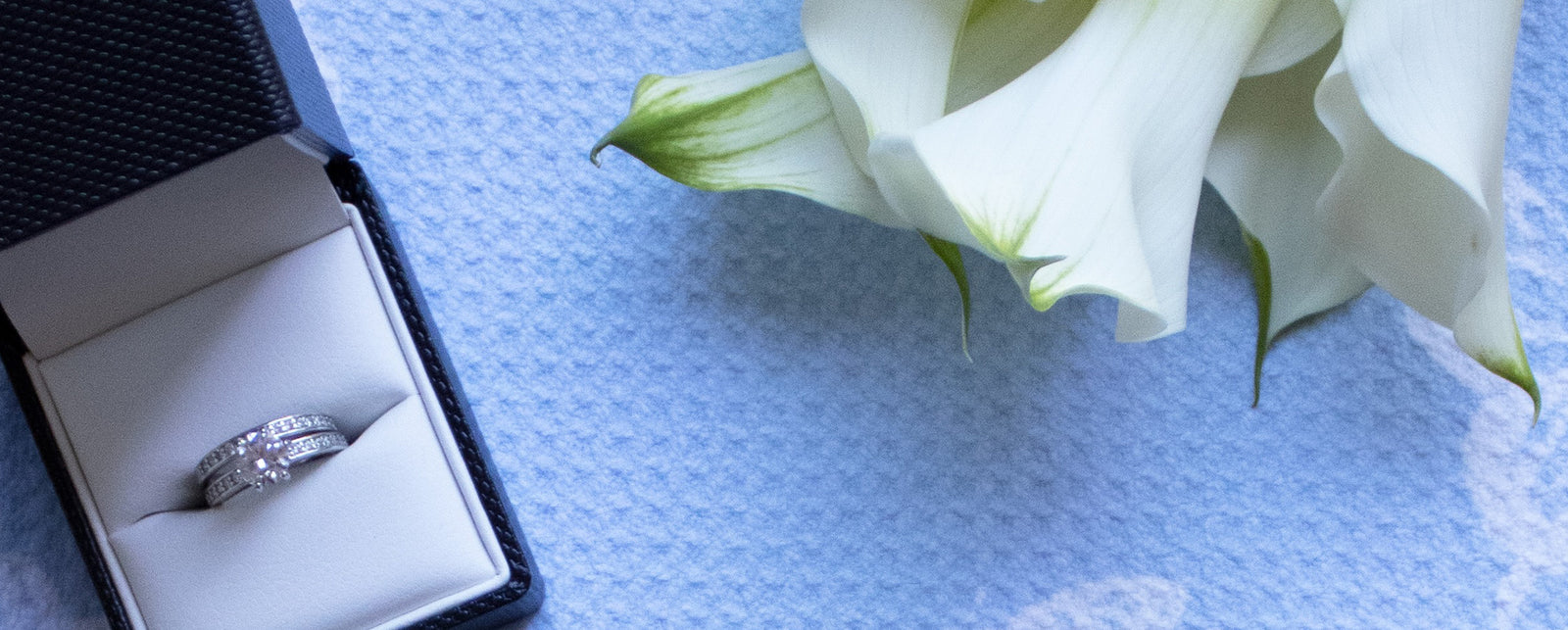 A diamond wedding ring in a black box sits on a light blue textured surface next to three white calla lilies.
