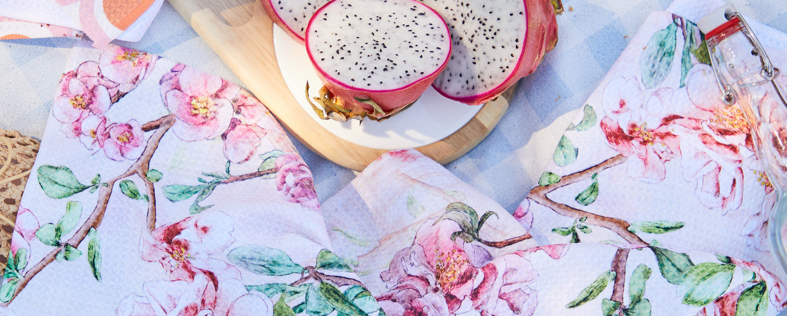 A picnic scene with a halved dragon fruit on a wooden board, floral-patterned napkins, and a blue-checked blanket.
